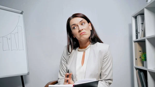 Заплутана бізнес-леді з блокнотом, що сидить біля фліпчарт в офісі — стокове фото