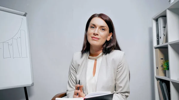 Businesswoman with pen and copybook looking at camera in office — Stock Photo