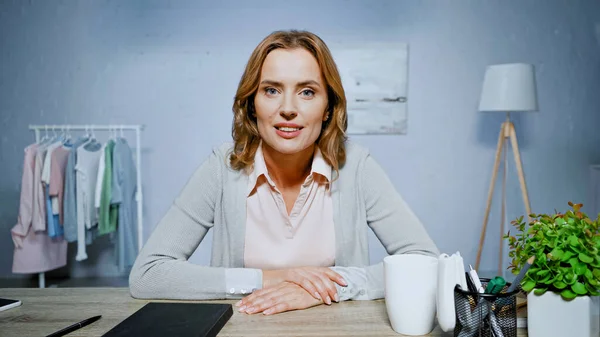 Lächelnde Frau blickt in die Kamera, während sie im Wohnzimmer am Tisch sitzt — Stockfoto