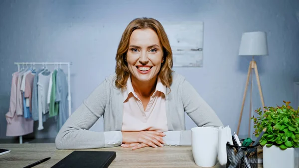 Positive woman smiling at camera near notebook, cup and stationery on table — Stock Photo