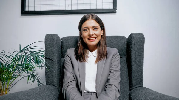 Brunette businesswoman in suit smiling at camera near plant in office — Stock Photo