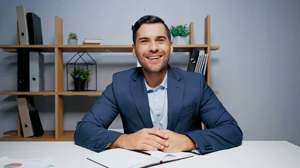 Smiling businessman looking at camera near notebook and papers on blurred foreground — Stock Photo