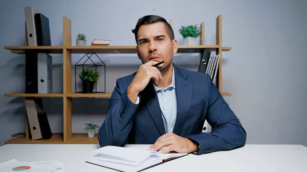 Pensativo hombre de negocios con pluma mirando a la cámara cerca de portátil - foto de stock