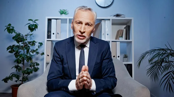 Elderly businessman with praying hands talking at camera during video call in office — Stock Photo