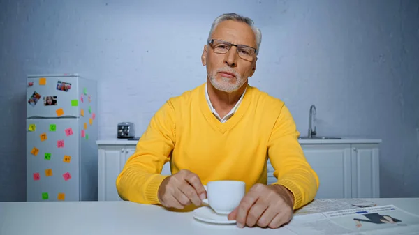 Uomo anziano guardando la fotocamera vicino giornale e tazza di caffè in primo piano sfocato — Foto stock