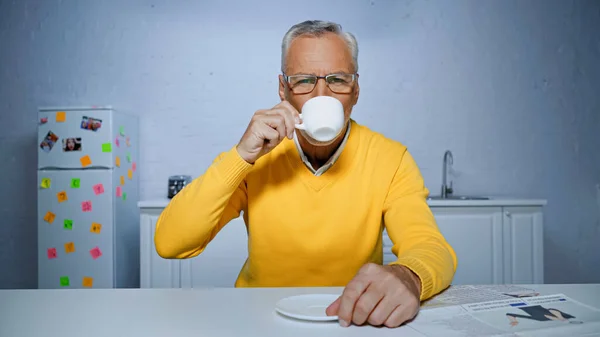 Uomo anziano in maglione giallo bere caffè vicino giornale in cucina — Foto stock