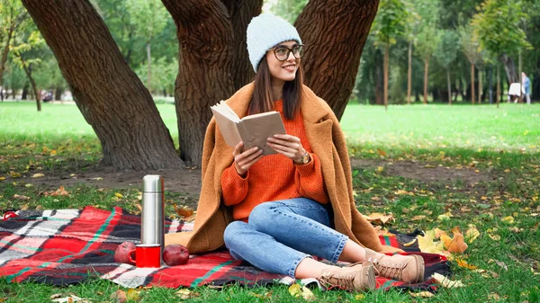 Mulher feliz olhando embora enquanto sentado no cobertor xadrez no parque com livro — Fotografia de Stock