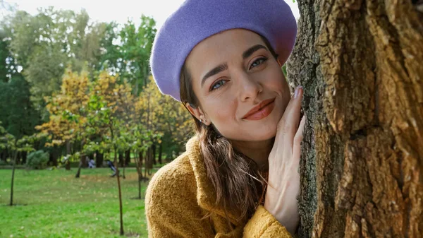 Веселая женщина в модном берете смотрит в камеру, опираясь на ствол дерева в парке — стоковое фото