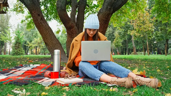 Jeune pigiste travaillant sur ordinateur portable tout en étant assis sur une couverture à carreaux sous l'arbre dans le parc — Photo de stock