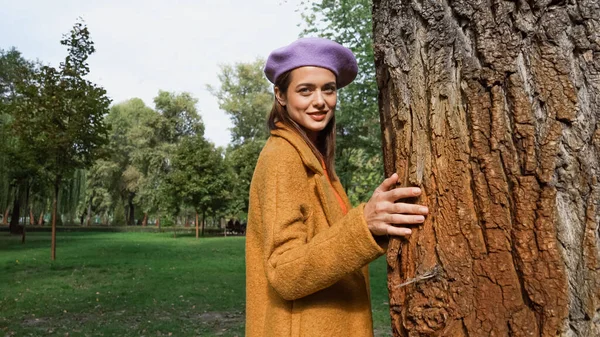Joven mujer de moda mirando a la cámara mientras está de pie cerca del tronco del árbol en el parque - foto de stock