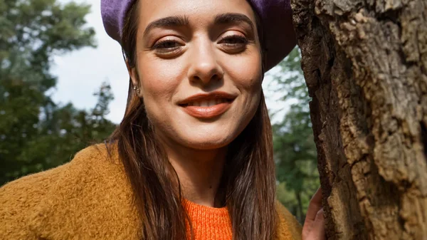 Portrait of cheerful woman smiling at camera near tree trunk in autumn park — Stock Photo