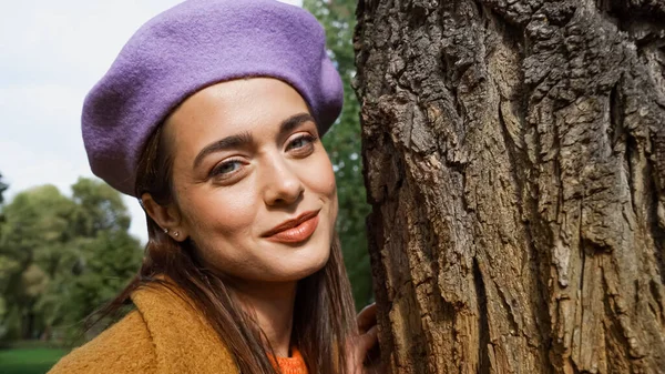 Mulher sorridente na boina elegante olhando para a câmera perto de tronco de árvore no parque de outono — Fotografia de Stock