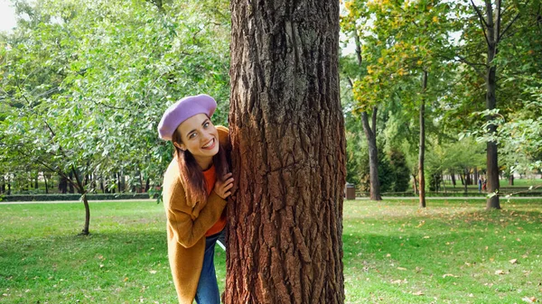 Excited woman hiding behind tree trunk in park while looking at camera — Stock Photo