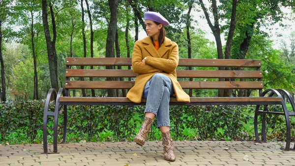 Trendy displeased woman looking away while sitting with crossed arms on bench in park — Stock Photo