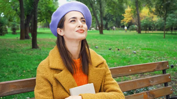 Donna felice che distoglie lo sguardo mentre si siede sulla panchina nel parco autunnale con libro — Stock Photo