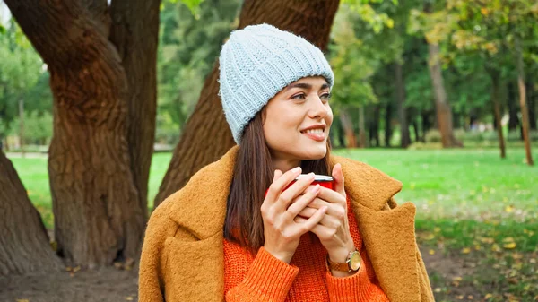 Happy woman in knitted beanie holding mug of warm drink in park — Stock Photo