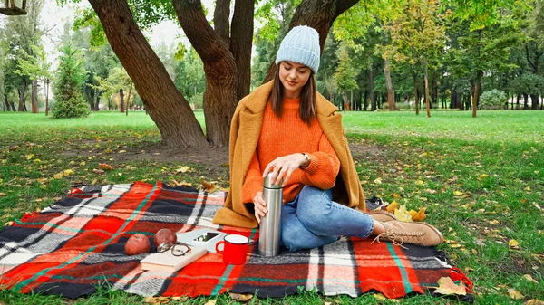 Жінка в стильному осінньому одязі відкриває термос під час пікніка в парку — стокове фото