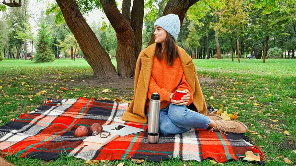 Улыбающаяся женщина в осеннем наряде смотрит в сторону, сидя на клетчатом одеяле в парке — стоковое фото