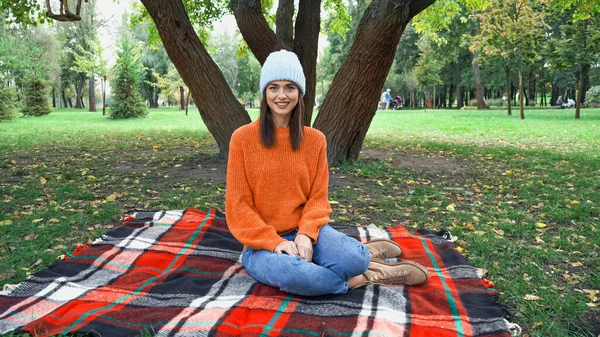 Heureuse femme élégante souriant à la caméra tout en étant assis sur une couverture à carreaux dans le parc — Photo de stock