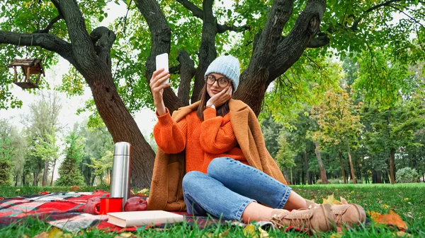 Femme souriante toucher le visage tout en prenant selfie pendant le pique-nique dans le parc — Photo de stock