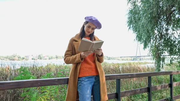 Junge Frau in trendiger Herbstkleidung liest Roman auf Brücke im Park — Stockfoto