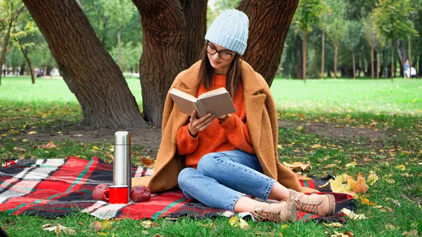 Стильная женщина в осенней одежде читает роман во время пикника в парке — стоковое фото