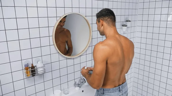 Joven hombre sin camisa sosteniendo desodorante cerca del espejo en el baño - foto de stock