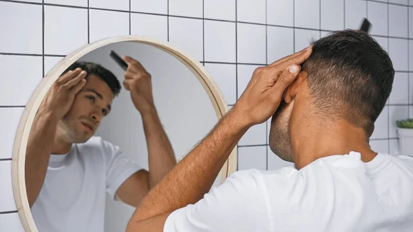 Junger Mann bürstet Haare mit Kamm in der Nähe von Spiegel im Badezimmer, verschwommener Hintergrund — Stockfoto