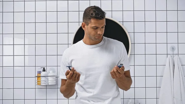 Man in white t-shirt holding safety razor and electric shaver in bathroom — Stock Photo