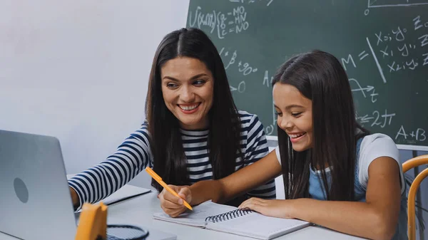 Mère souriant et regardant ordinateur portable près de fille heureuse faire des devoirs — Photo de stock