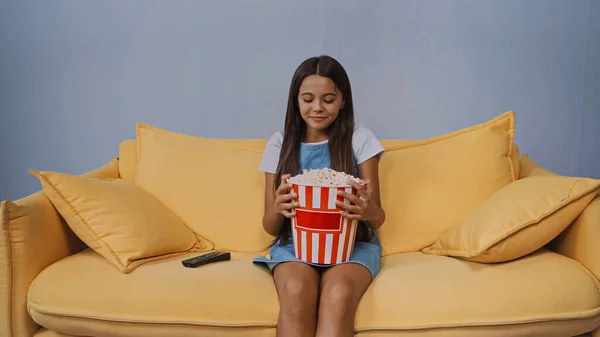 Happy kid looking at bucket with popcorn while sitting on sofa — Stock Photo