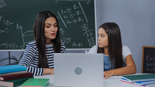 Madre e hija haciendo la tarea cerca de la computadora portátil y cuadernos en la mesa - foto de stock