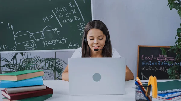 Heureux écolier dans casque regardant ordinateur portable tout en étudiant en ligne à la maison — Photo de stock