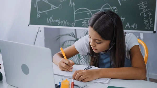 Школярка в гарнітурі пише в блокноті біля ноутбука на столі — Stock Photo