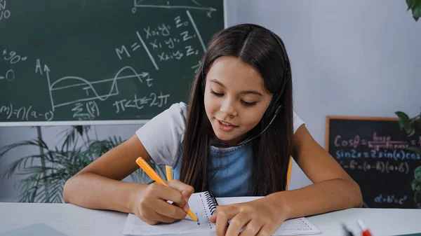 Schulkind in Headset schreibt in Notizbuch auf Schreibtisch — Stockfoto