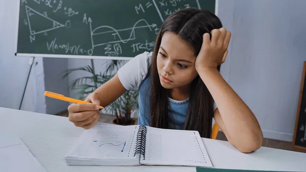 Niño disgustado sosteniendo la pluma y mirando el cuaderno con gráfico - foto de stock