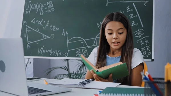 Kind liest Buch neben Laptop und Notizbuch auf Schreibtisch vor — Stockfoto