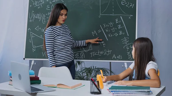 Madre señalando con la mano en pizarra con fórmulas matemáticas cerca de la hija - foto de stock