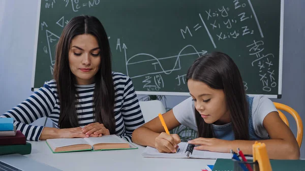 Mère regardant livre près de fille écriture sur ordinateur portable tout en faisant des devoirs — Photo de stock