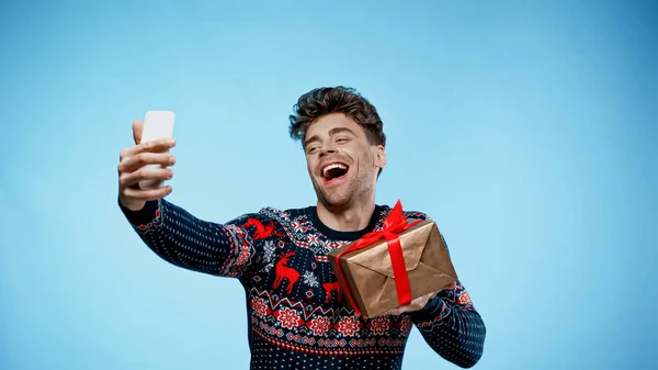 Веселый мужчина с подарочной коробкой делает селфи на синем фоне — стоковое фото