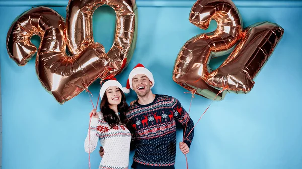 Happy couple in santa hats and sweaters holding balloons in shape of 2021 on blue background — Stock Photo