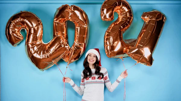 Smiling woman in santa hat holding balloons in shape of 2021 numbers on blue background — Stock Photo
