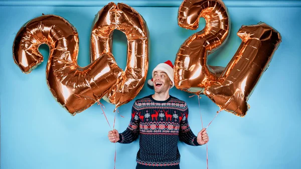 Homem incrível em suéter e chapéu de Papai Noel segurando balões em forma de 2021 números no fundo azul — Fotografia de Stock
