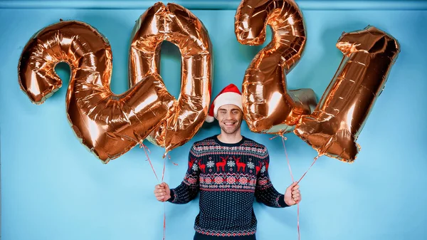 Homem sorridente em suéter e chapéu vermelho de Papai Noel segurando balões dourados em forma de 2021 números no fundo azul — Fotografia de Stock