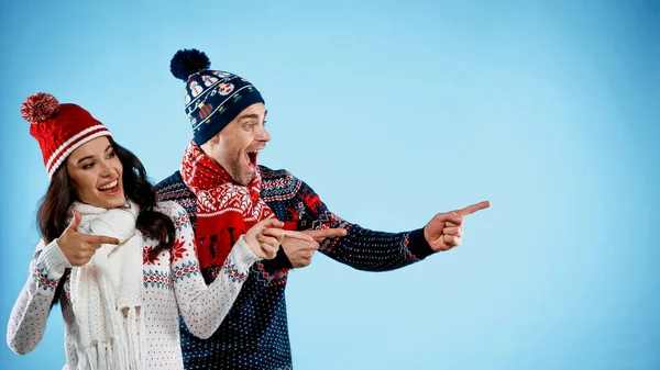 Cheerful couple in hats and sweaters pointing with fingers on blue background — Stock Photo