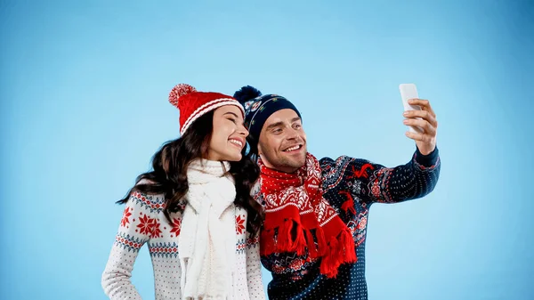 Happy couple in hats, sweaters and scarfs taking selfie on blue background — Stock Photo