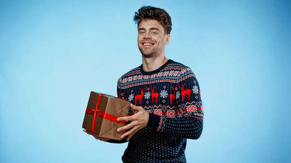 Усміхнений чоловік у теплому светрі тримає подарунок на синьому фоні — стокове фото