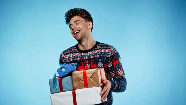 Cheerful man in sweater holding presents on blue background — Stock Photo