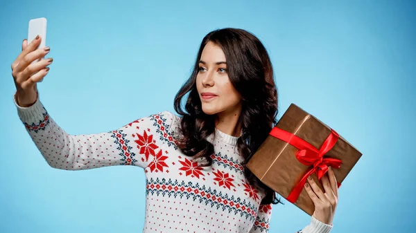 Young woman in warm sweater taking selfie and holding present on blue background — Stock Photo