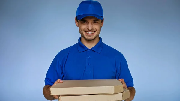 Alegre repartidor hombre sosteniendo cajas de pizza mientras sonríe a la cámara aislado en azul - foto de stock